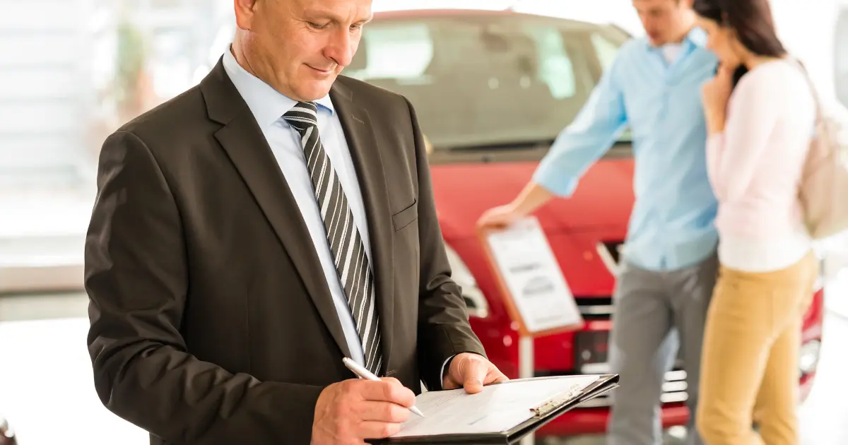 Car dealer reviewing purchase paperwork while buyers inspect a vehicle