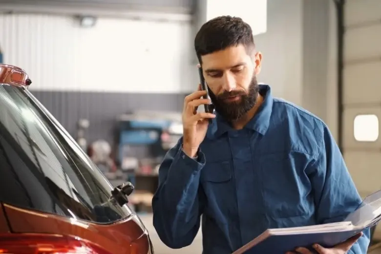 Mechanic speaking on the phone while reviewing repair paperwork beside a car